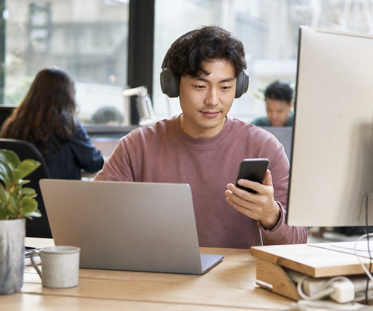A web designer testing a page layout on a laptop and a phone while wearing headphones